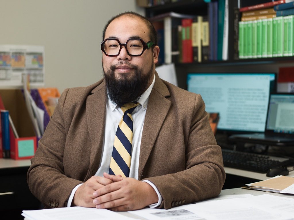 An Asian-Canadian man who is balding and has a pointed black beard sits at his desk. He has dark glasses and is wearing a brown jacket, tie, and white shirt. Behind hi